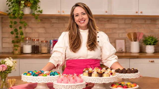 Veri Fragoso sorrindo na cozinha, atrás de uma mesa com bombons e doces finos em boleiras